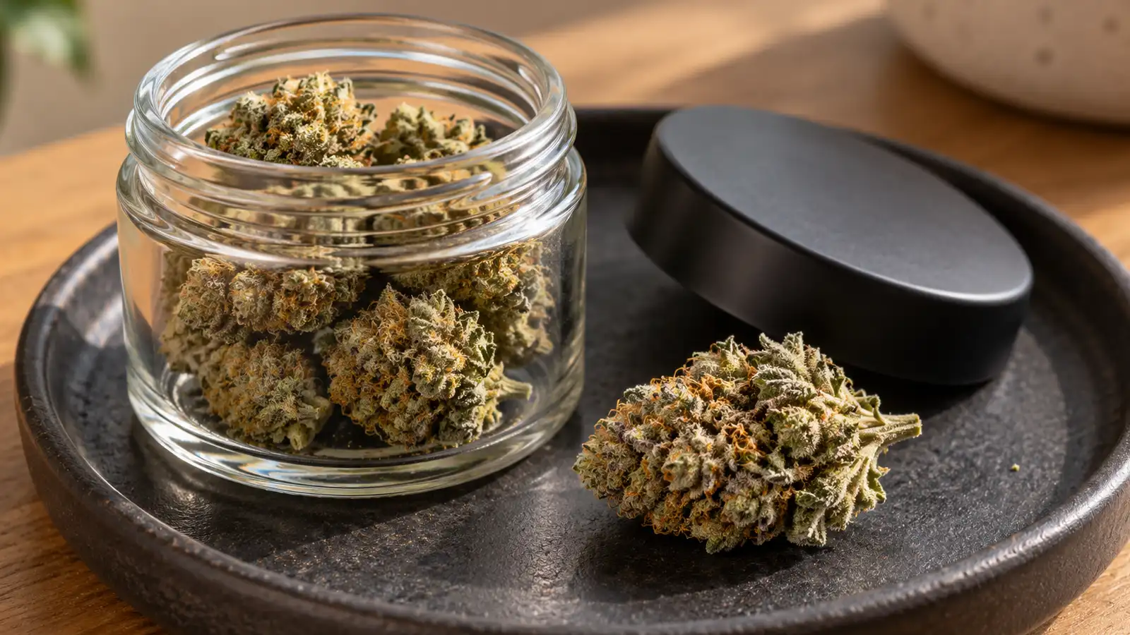 Dried cannabis flowers sit in a plain glass jar and on a dark ceramic tray.
