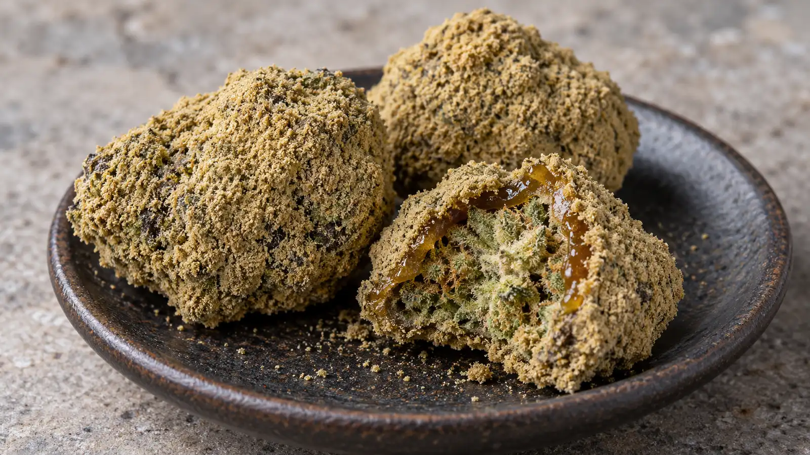 Kief-coated cannabis moonrocks sit on a dark saucer, with one broken open to reveal the flower inside.
