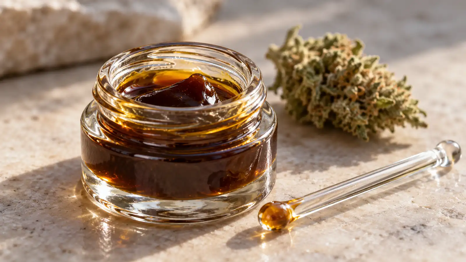 Dark amber cannabis oil fills a small glass jar beside a glass rod and a dried flower.