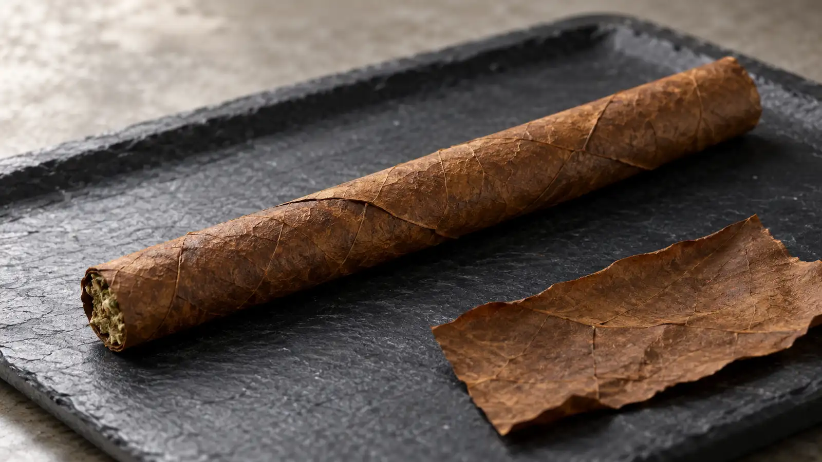 An unlit blunt in a brown tobacco leaf wrapper rests on a slate tray beside a piece of wrapper.