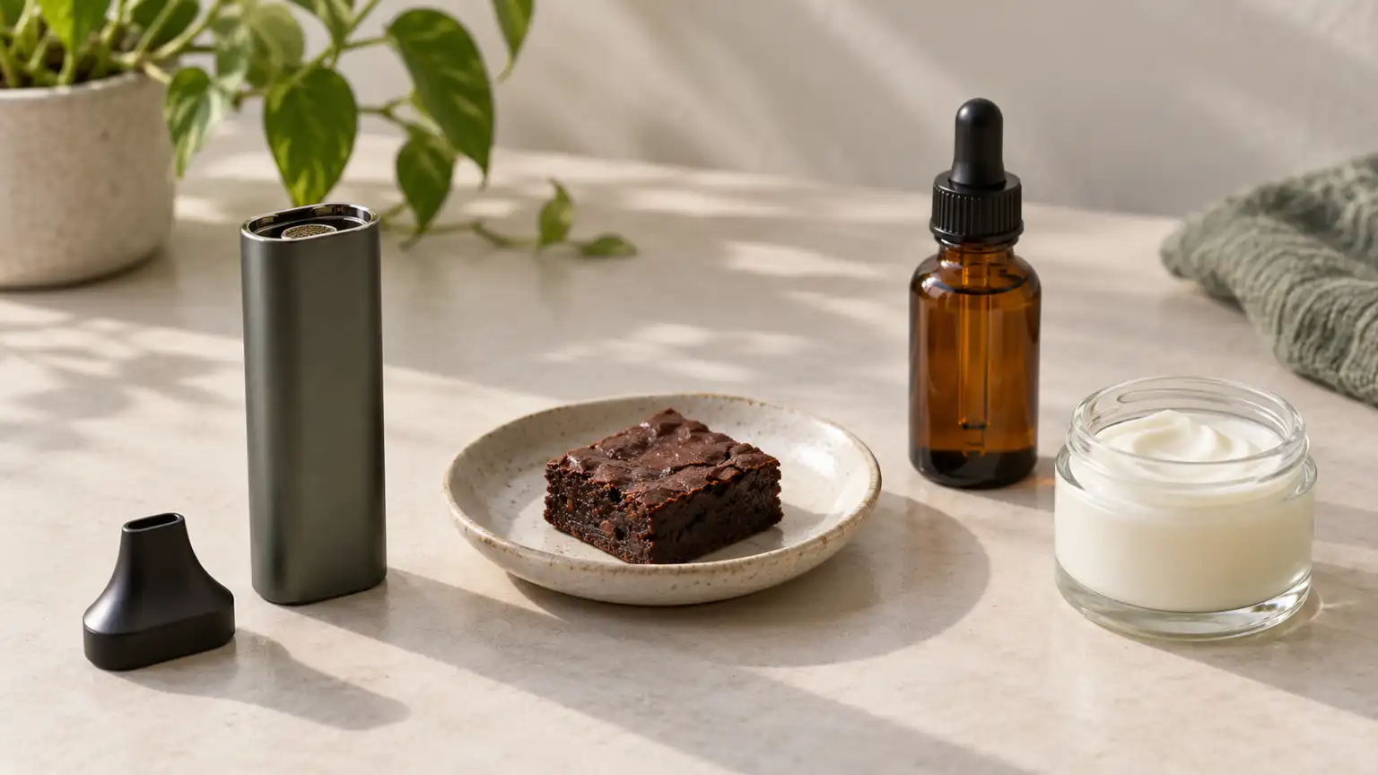 A vaporizer, brownie, amber dropper bottle, and cream jar sit on a pale tabletop.