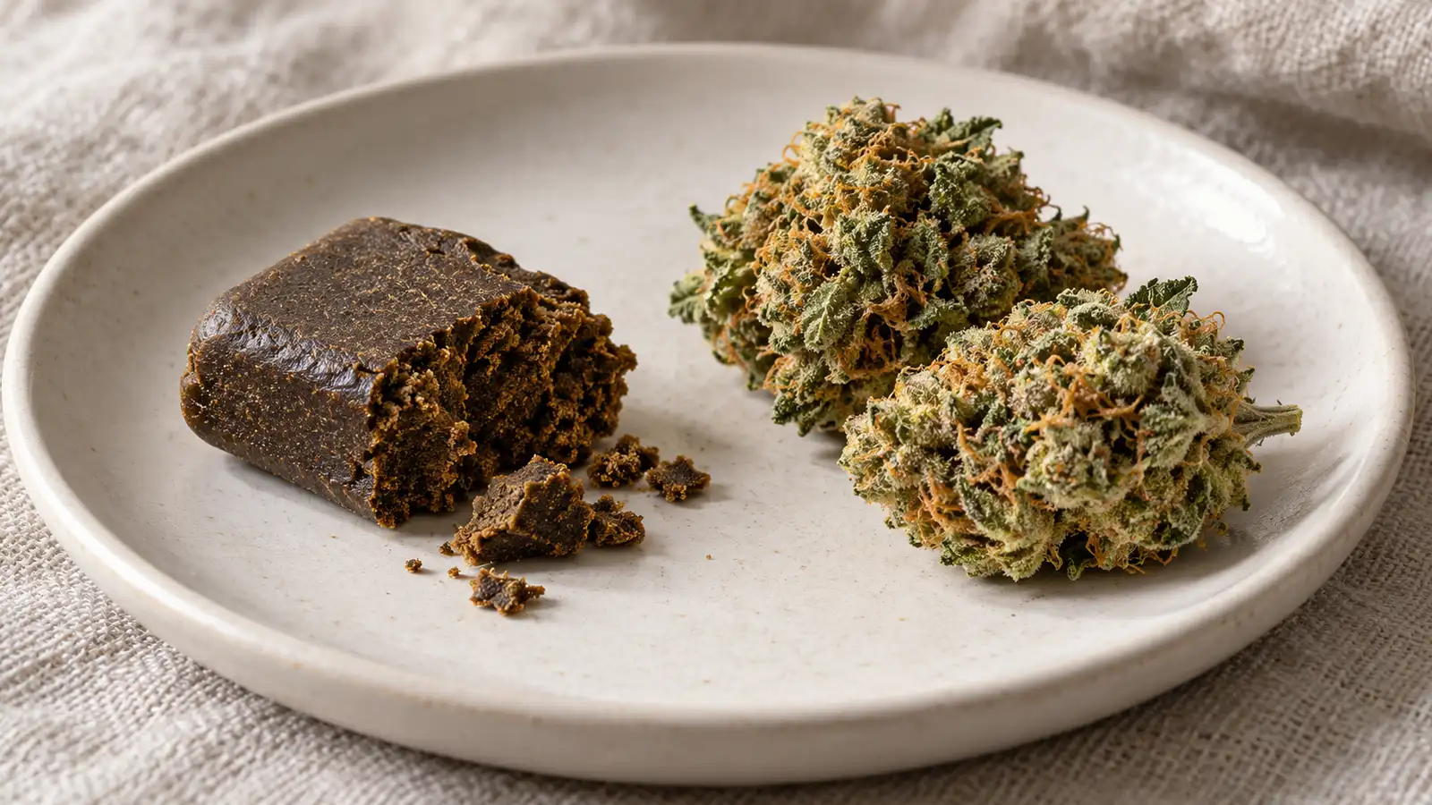 A crumbly piece of brown hashish lies beside dried cannabis flowers on a ceramic plate.