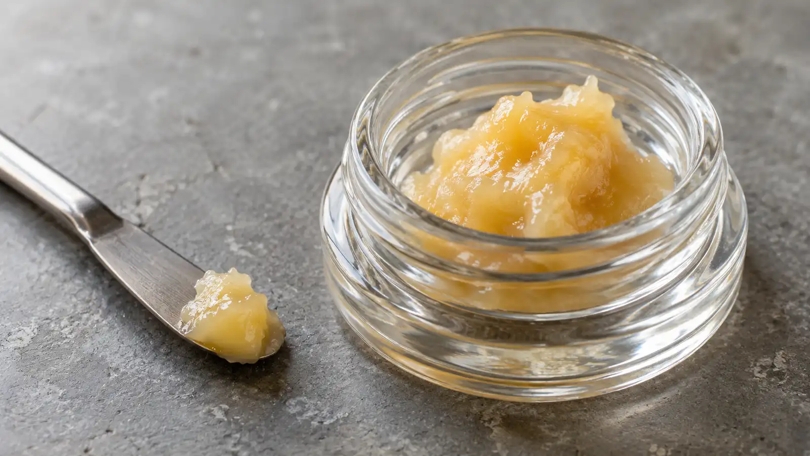 Pale golden live rosin sits in a small glass jar beside a stainless steel spatula.